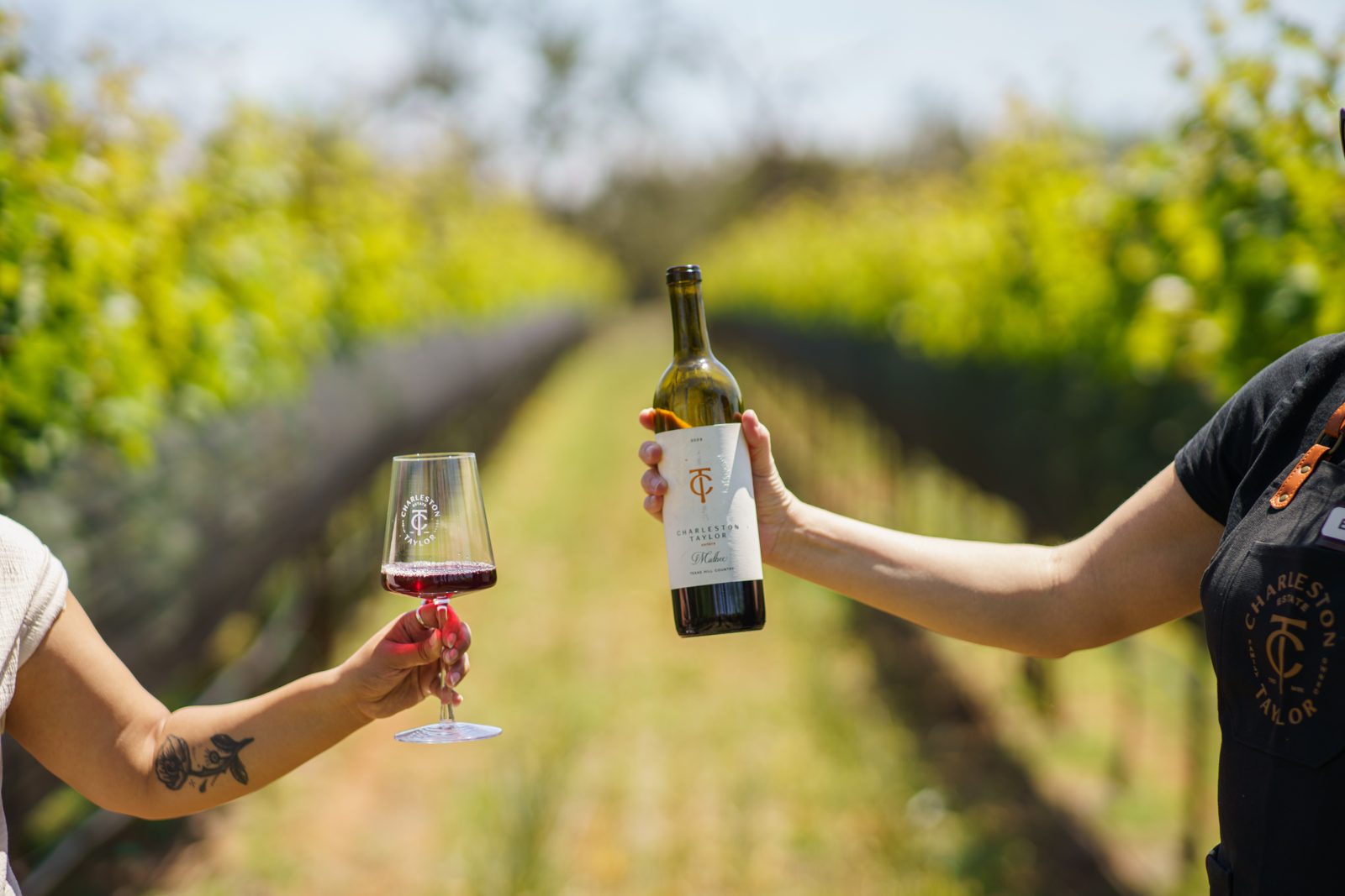 A glass of red and a bottle held up between vineyard rows in the Texas Hill Country